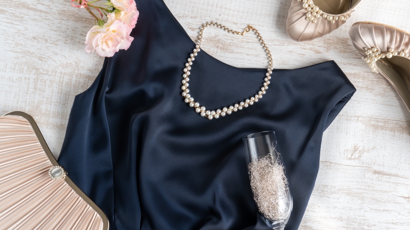 パーティードレスの周りにパールのネックレスやパンプス、バッグやシャンパングラス、お花が置かれている