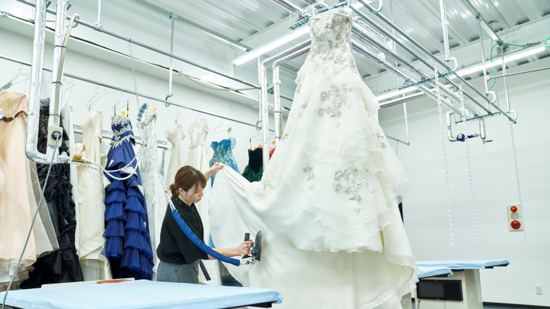 高い位置から吊り下げた大きなウエディングドレスのプレスをするディアハートのスタッフ